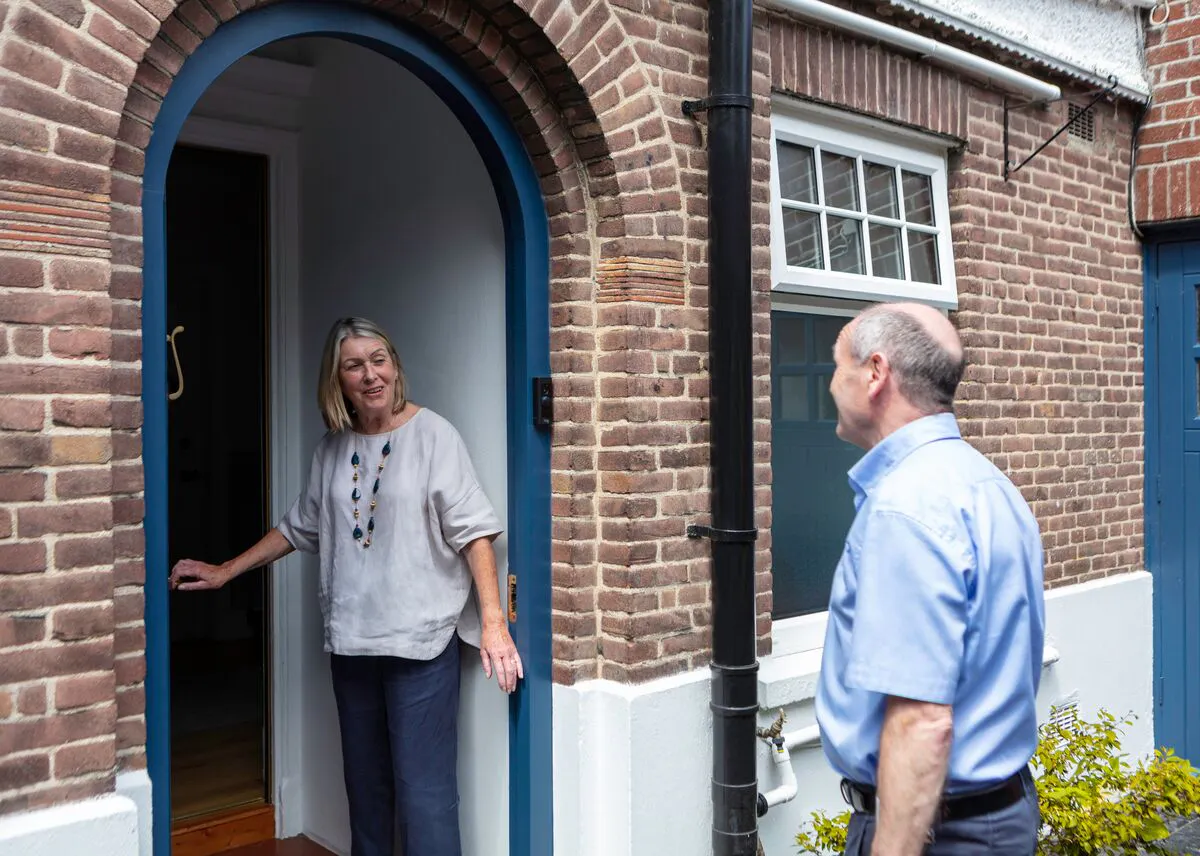 Exterior of house with curved brick porch with blue trim, a woman welcomes a man at the doorstep.
