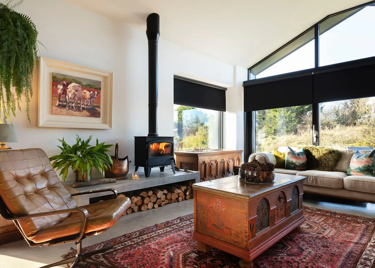 Gable end living room in cream with windows dressed in black roller blinds, lit stove, pale beige sofa in front of window, leather chair, wooden chest.