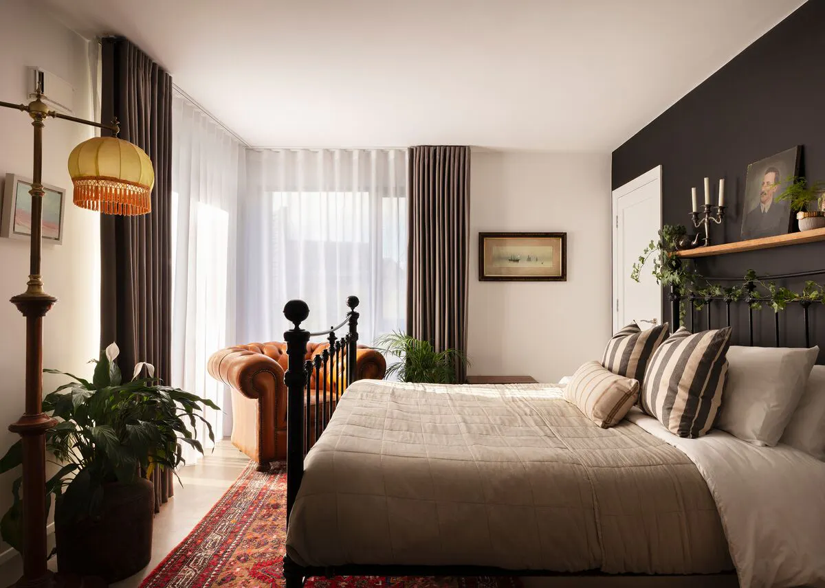 Neutral bedroom with one midnight blue wall behind bed, corner windows dressed in white voile curtains, closed, under mid brown curtains open, wrought iron style bed with pale brown bedding, tan chesterfield chair by windows, fringed lamps on stand.