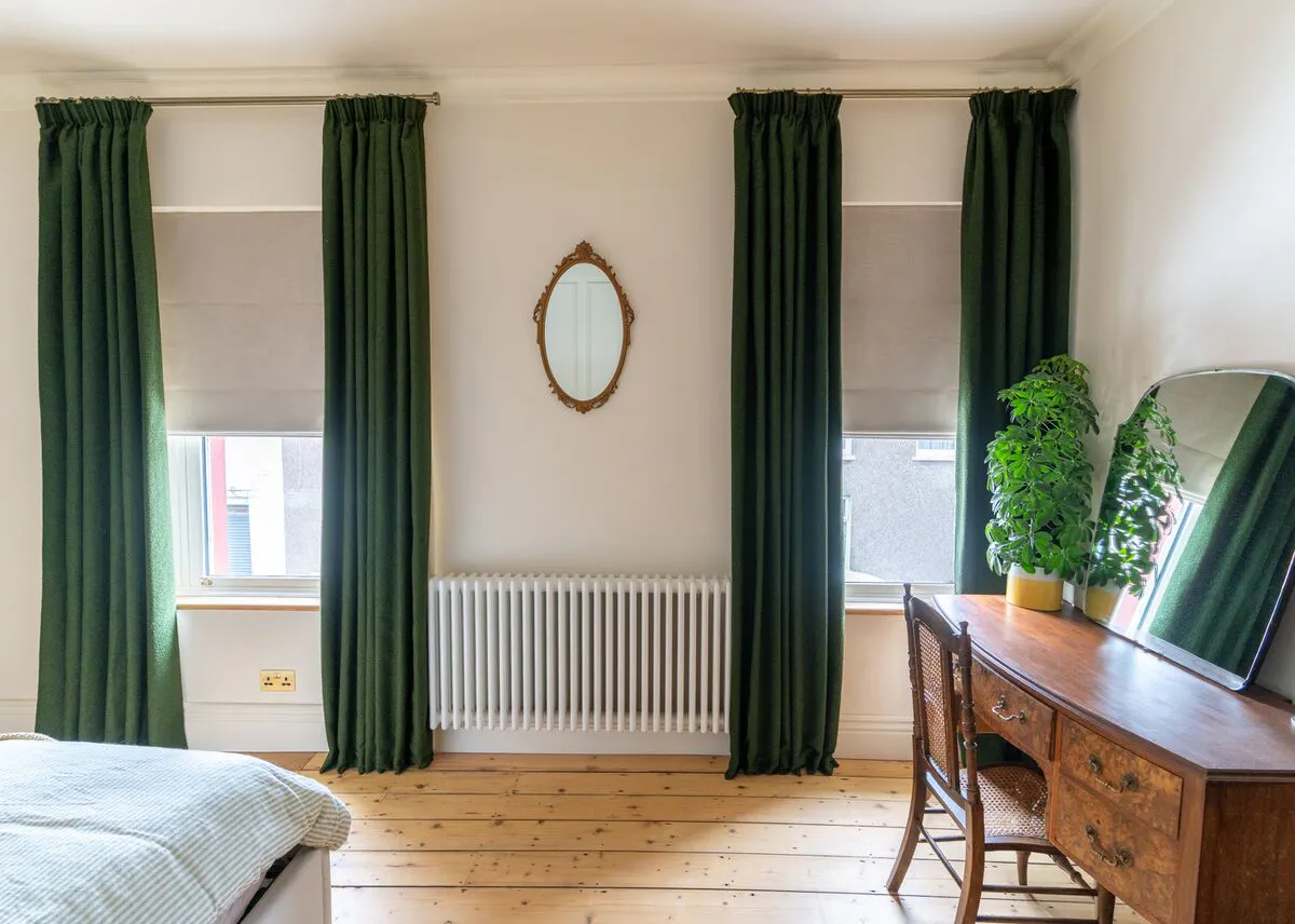 Neutral bedroom with two windows each dressed in putty coloured roman blinds lowered halfway, under forest green curtains, open, wooden floor, wooden dressing table. 
