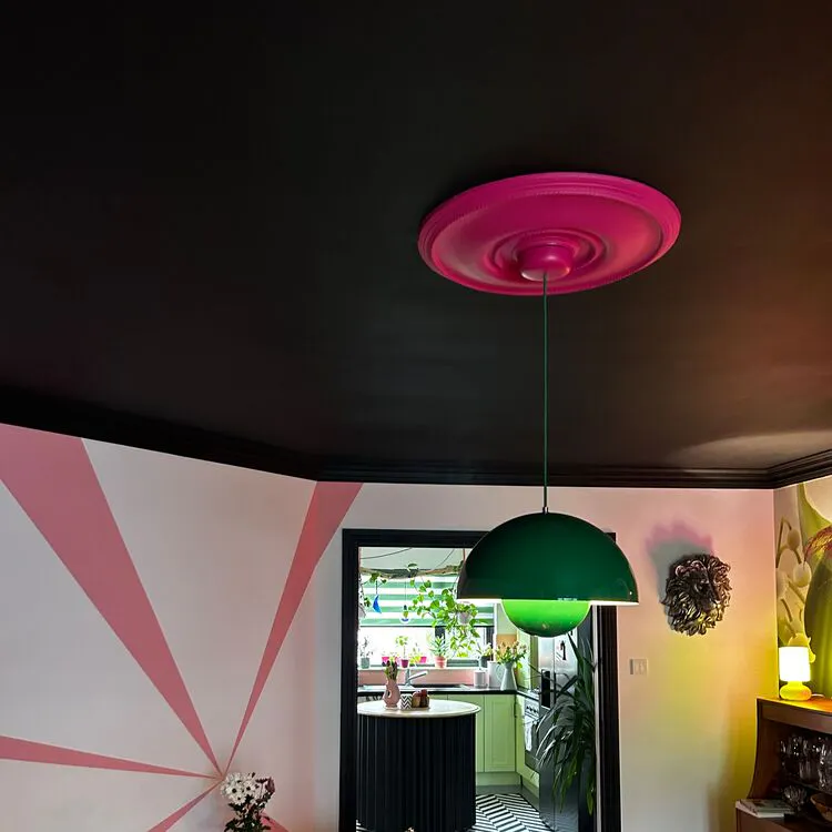 Living room with black ceiling, deep pink ceiling rose, green lamp shade, sofa with multiple cushions in different colours.