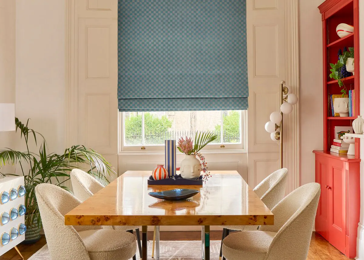 Cream dining room with window dressed in blue checkerboard Roman blind lowered halfway, wooden dining table with cream chairs, terracotta dresser.