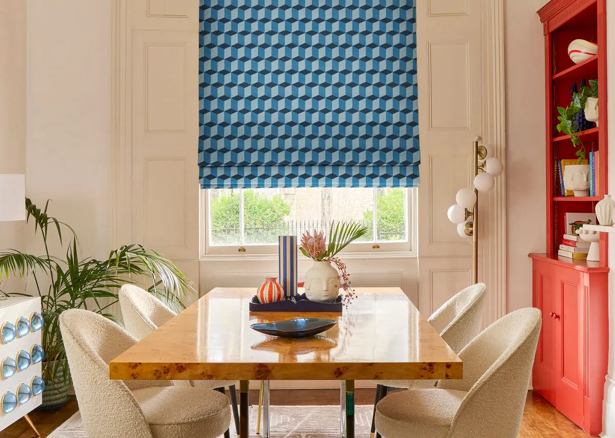 A blue geometric Roman blind covers a large window in a bright dining room with neutral chairs and warm accents. 