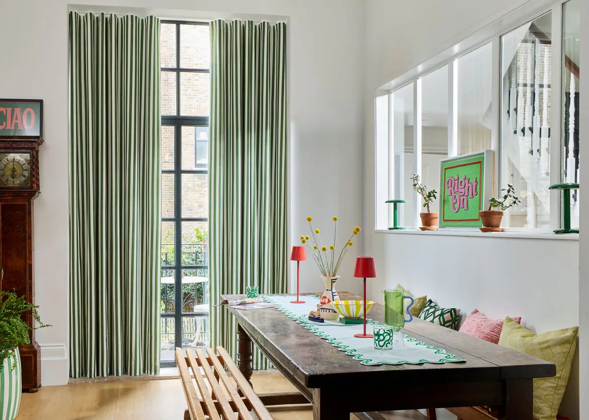 Green striped curtains frame a tall black‑framed window beside a bright dining area with colourful table décor.