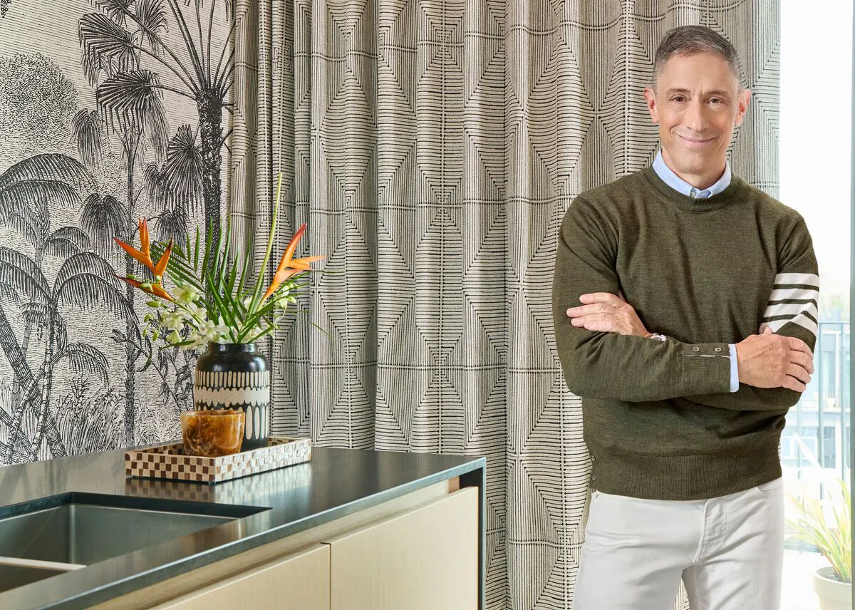 Man stands beside a kitchen island, with patterned curtains drawn across a bright window, adding texture and softness to the modern space. 