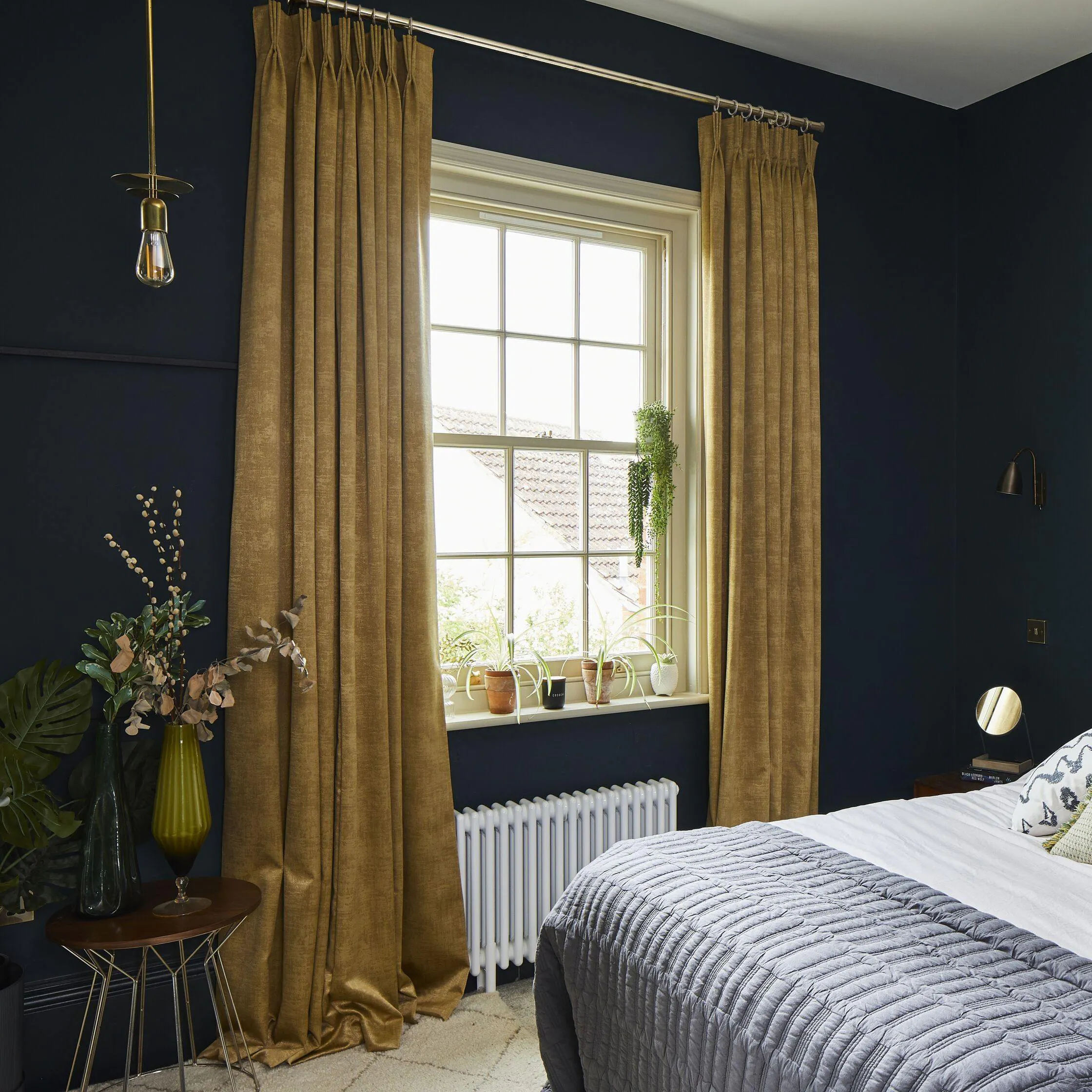 Mustard curtains in a bedroom. There is a bed on the left hand size that takes up the room. There is plotted plants in the room by the window and bed.