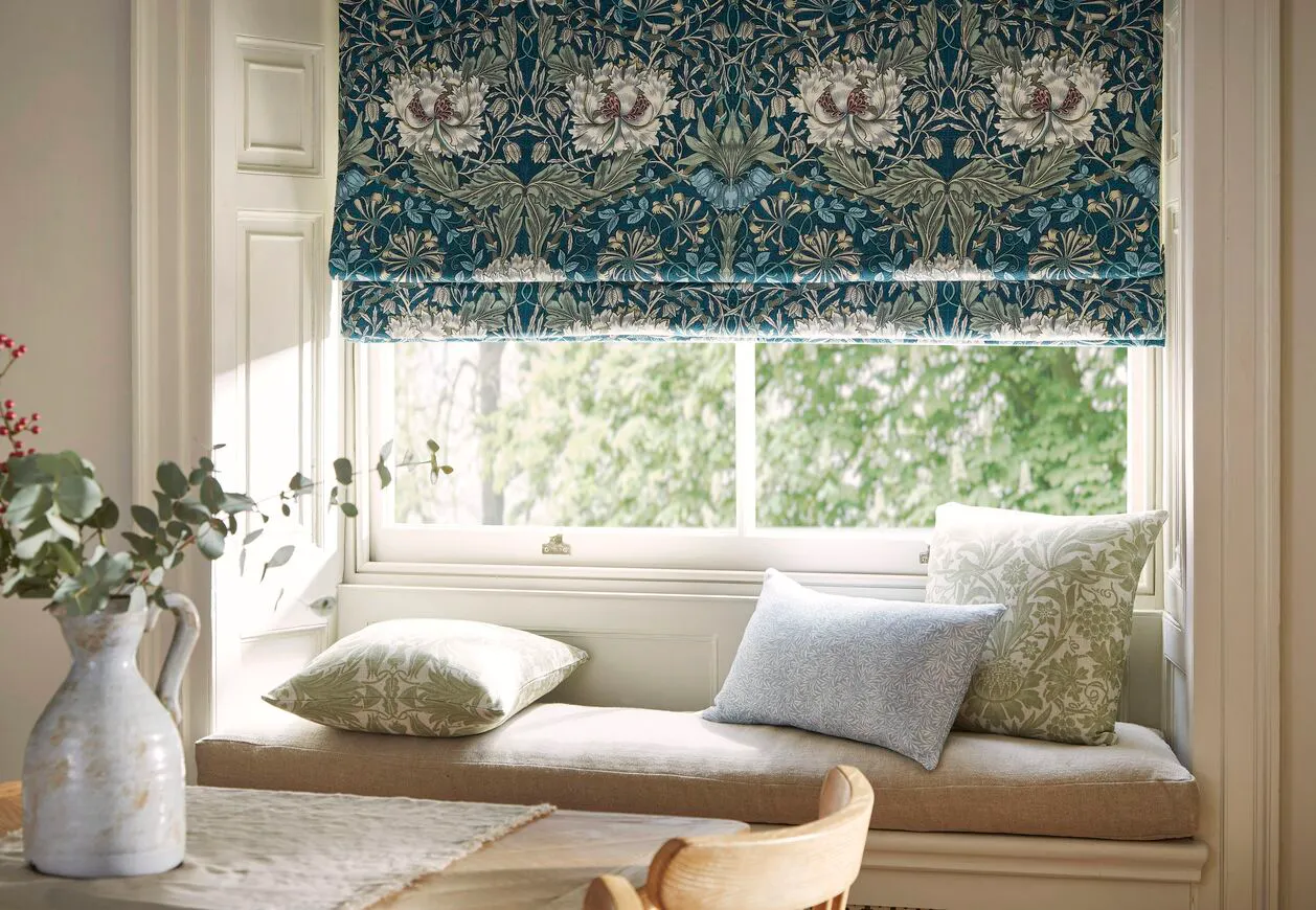 A patterned Roman blind sits neatly above a bright window, with soft daylight filtering onto a built-in window seat and cushions.