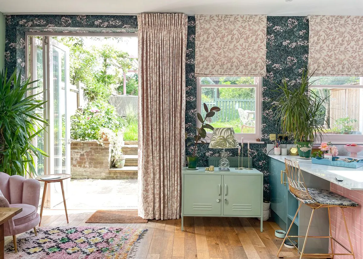 Patterned Roman blinds and curtain brighten a colourful kitchen, framing leafy garden views and filling the space with natural light.