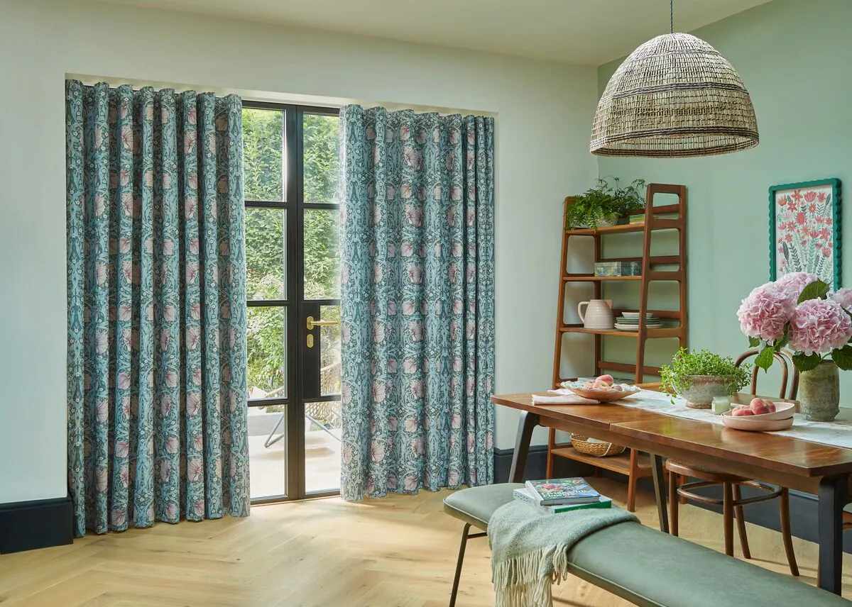 Patterned curtains frame tall, glazed doors, letting daylight into a relaxed dining area with a wooden table, greenery and soft green accents.