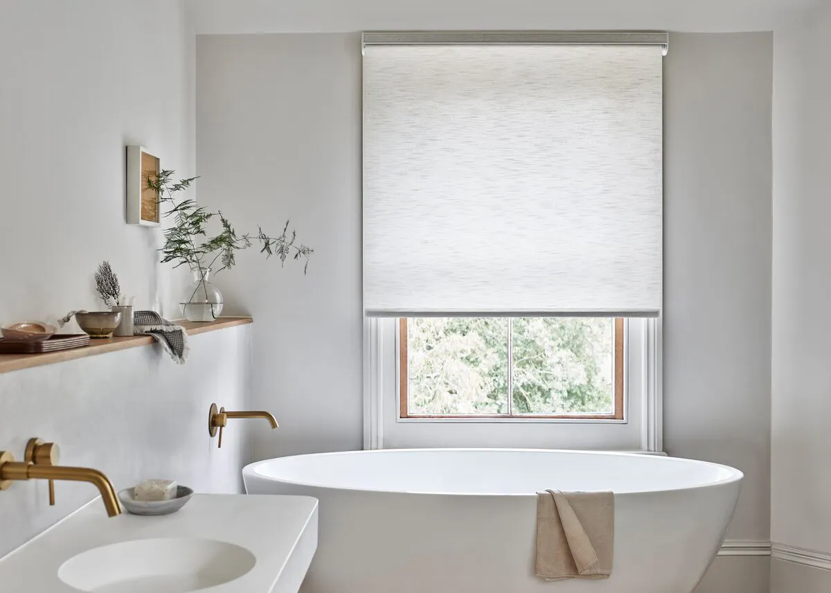 A light-filled bathroom window with a pale Roller blind, set above a freestanding bath and framed by calm, neutral walls. 