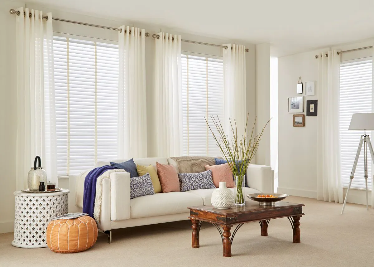 Tall living room windows with white blinds and voile curtains, filling the space with soft daylight behind a neutral sofa and layered décor.