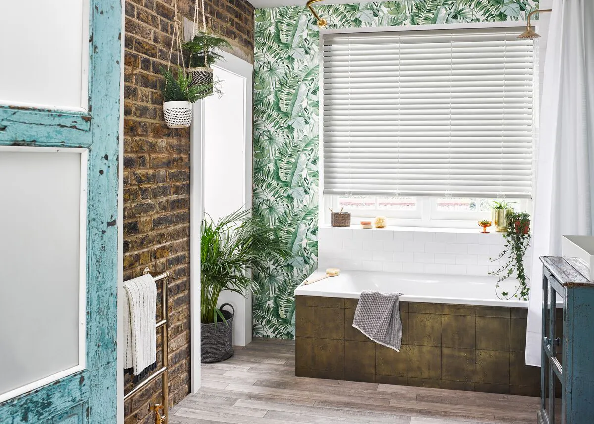 Bathroom with brick-look wall to side, window wall in green botanical print and window dressed in white faux wood blind, lowered three quarters, bath with wood effect panel, wood effect floor, multiple fern-type plants.