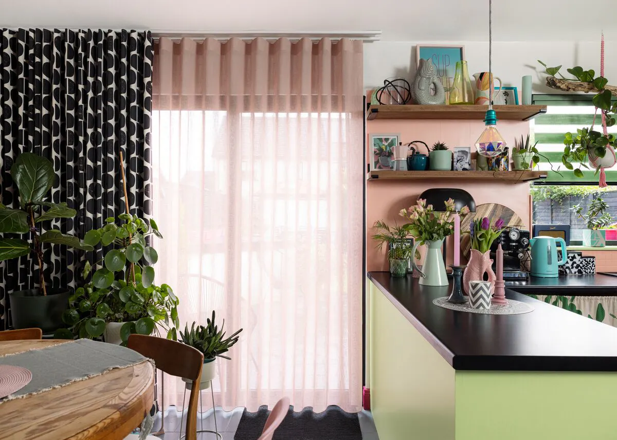 Kitchen-diner with large glazed doors framed by soft pink sheer curtains, allowing daylight to filter into a colourful, plant-filled space.