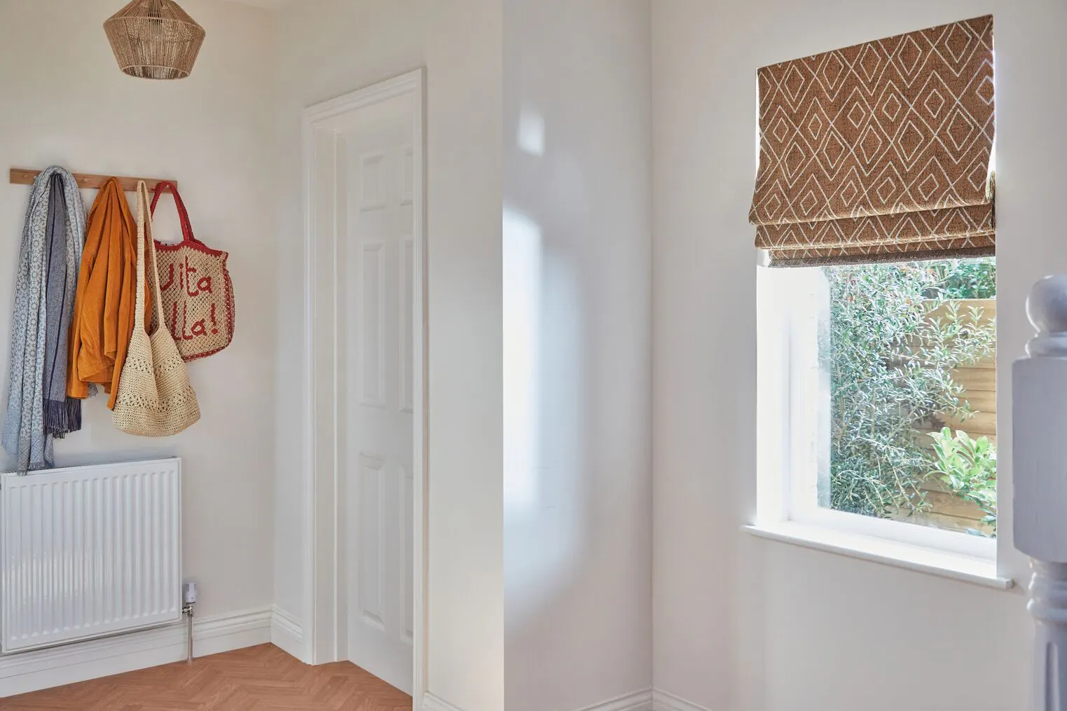 Bright hallway window dressed with a patterned brown Roman blind, filtering daylight and framing leafy garden views outside.