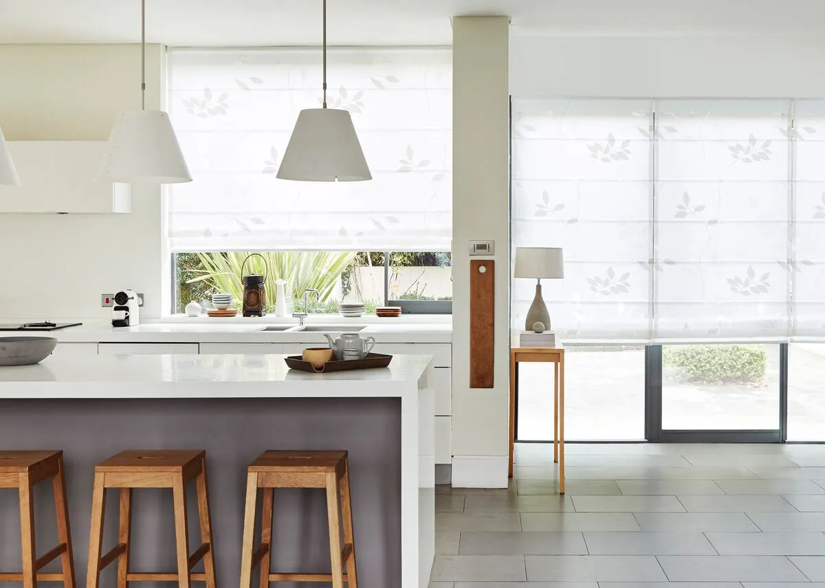Bright kitchen with wide glazing dressed in Voile Romanblinds, flooding the space with light above an island and wooden stools.