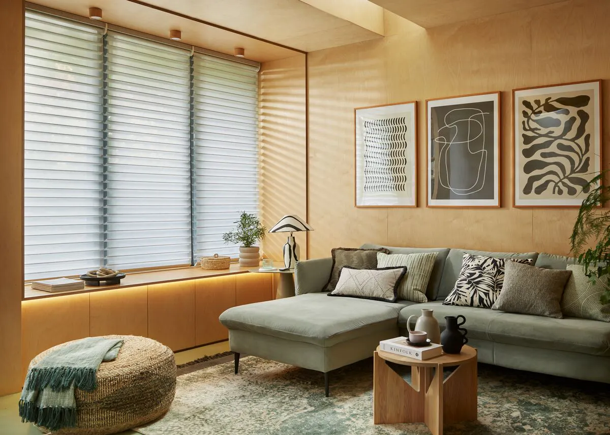 Cosy living room with a wide window fitted with a Triple Shade blind, softly filtering daylight across a green corner sofa and warm wooden finishes.