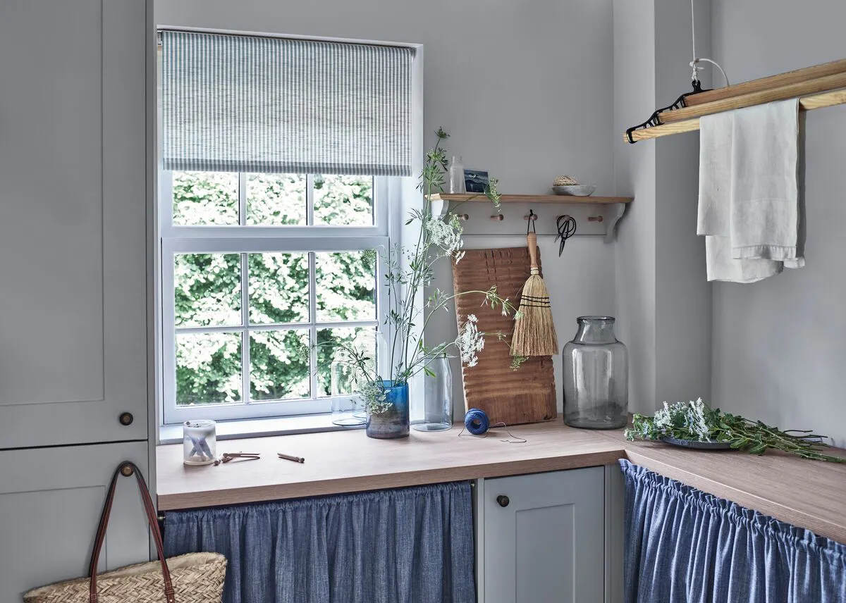 Utility room with a window dressed in a soft blue blind, letting daylight fall over a wooden worktop, open shelves and skirted cupboards.