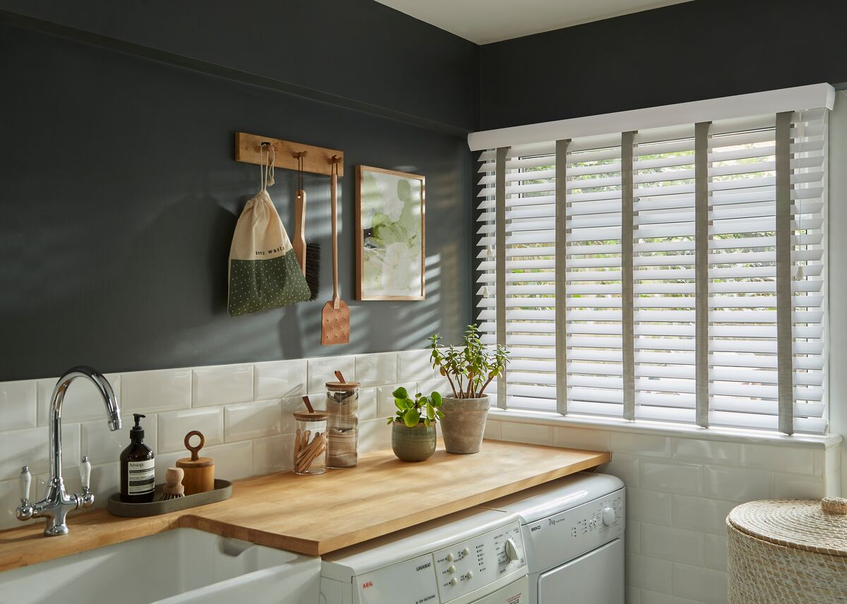 Utility room with a window fitted with a white faux wood blind, bringing soft light onto wooden worktops, a Belfast sink and neatly stacked laundry appliances.