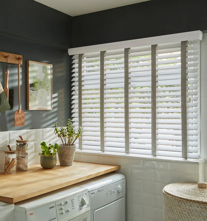 Utility room with a window fitted with a white faux wood blind, bringing soft light onto wooden worktops, a Belfast sink and neatly stacked laundry appliances.