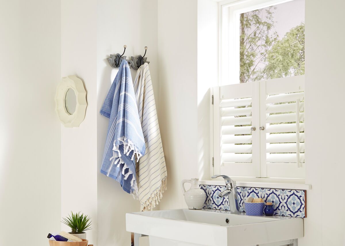 Utility sink area with a small window fitted with a white shutter, letting in natural light above blue patterned tiles and a simple basin.