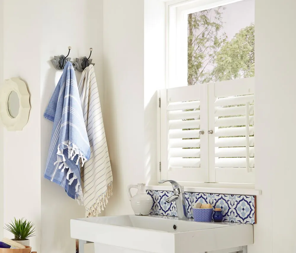Utility sink area with a small window fitted with a white shutter, letting in natural light above blue patterned tiles and a simple basin.