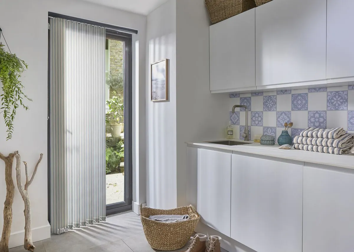 Utility room with a glazed door fitted with Vertical blinds, letting in garden light beside white cabinets, tiled splashback and woven baskets.