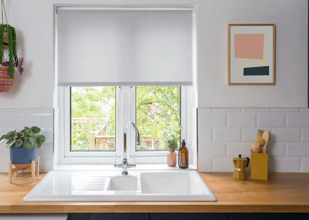 Kitchen with a window dressed in a Roller blind above a white sink, letting natural light onto wooden worktops, plants and tiled splashback.
