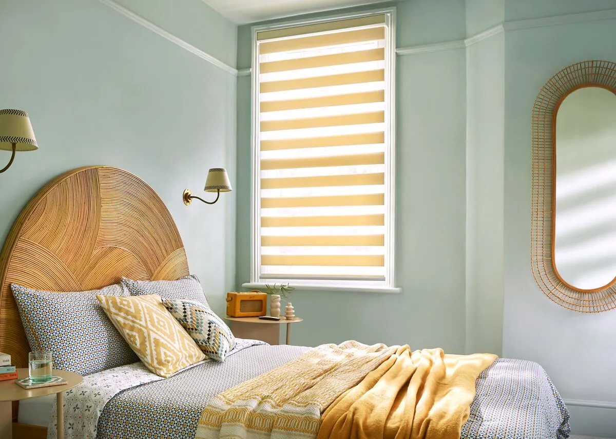 Bedroom featuring a tall window fitted with a Day and Night blind, softly filtering daylight above a neatly styled bed in calming pastel tones.