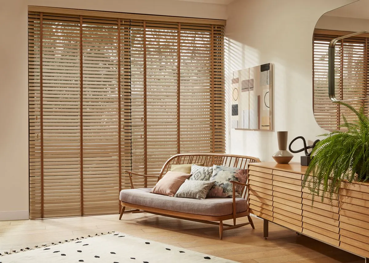 Living room with a wide window covered by a Wooden blind, casting warm filtered light across a rattan sofa and sleek sideboard.