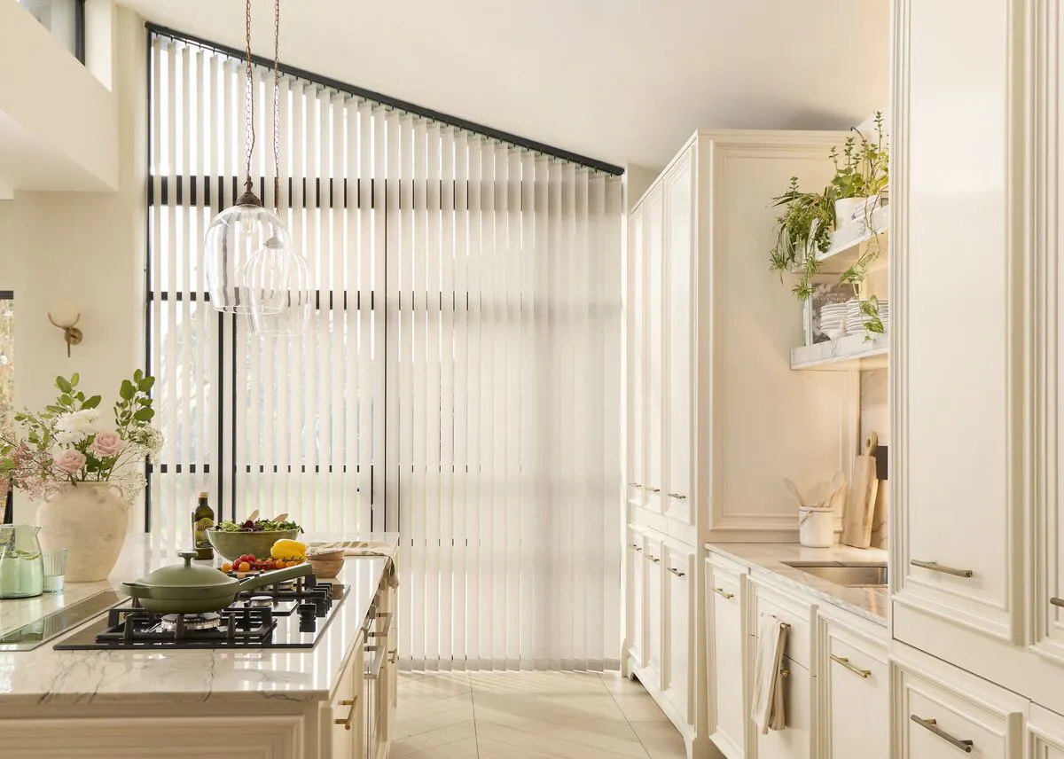Kitchen with a large window dressed in a Vertical blind, allowing soft daylight to filter across pale cabinetry, a central island and hanging glass pendants.