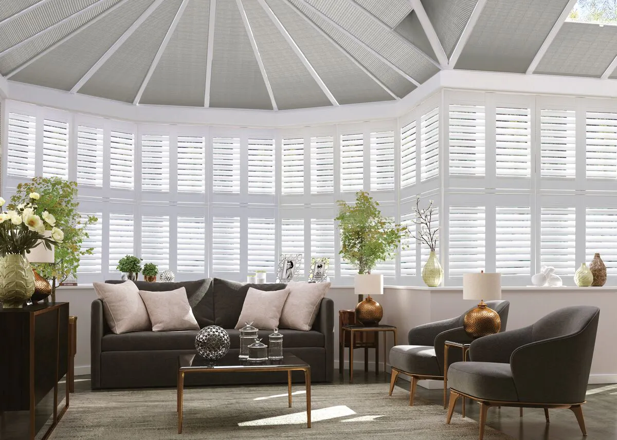 Elegant conservatory seating area with tall windows fitted with crisp white shutters and roof glazing softened by subtle blind panels, flooding the room with light.