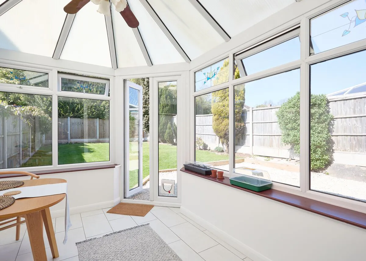 White conservatory with wide glazed windows and roof panels, letting daylight flood a dining area and framing a clear view of the garden outside.