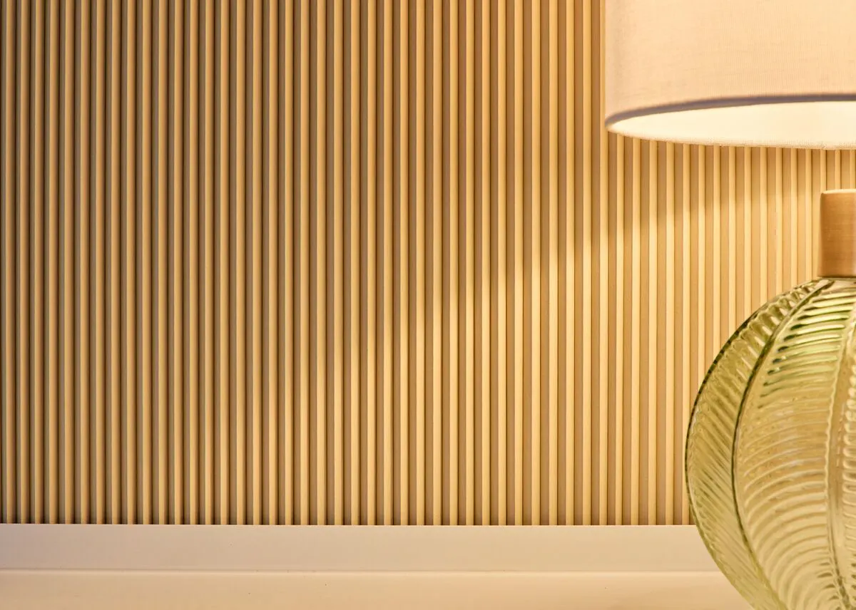 A close up of a warm-toned Blind Screen with a large ribbed green glass table lamp in the foreground.