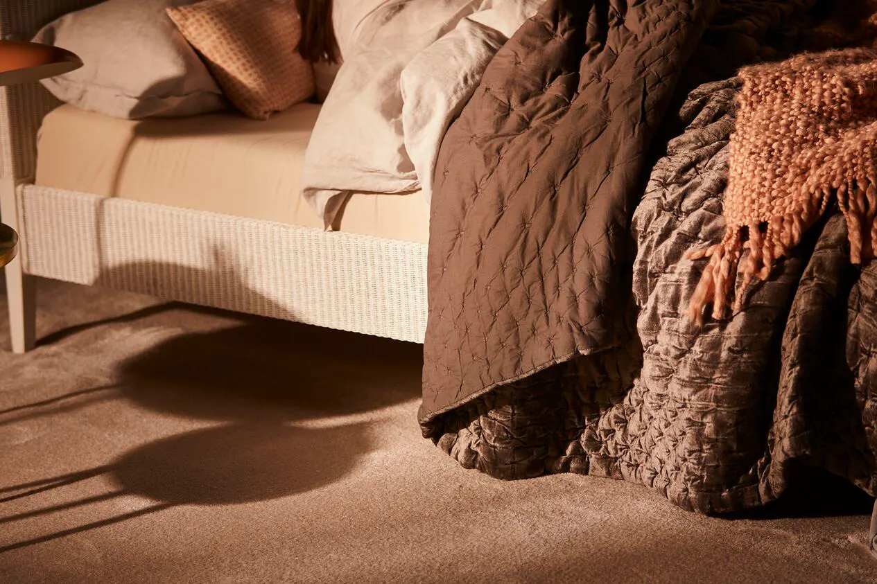A cosy close-up of a white bed frame on a deep carpet. The covers are pulled back and the bed is layered with pillows, a thick duvet and rich brown throws. 