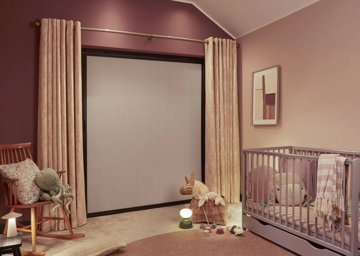 A rose-toned nursery with a total blackout blind framed by blush velvet curtains, with a grey cot, rocking chair and scattered toys.