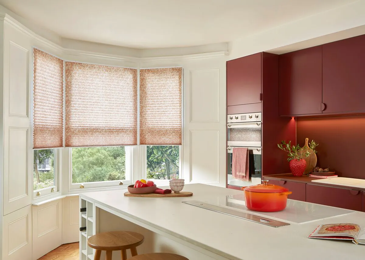Bay window with a light blind softening daylight, overlooking greenery and brightening a modern kitchen with warm red cabinetry and island seating.
