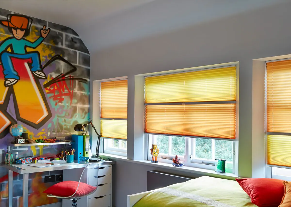 Row of windows fitted with orange and yellow blinds, casting warm light into a colourful bedroom with mural artwork and a compact workspace.