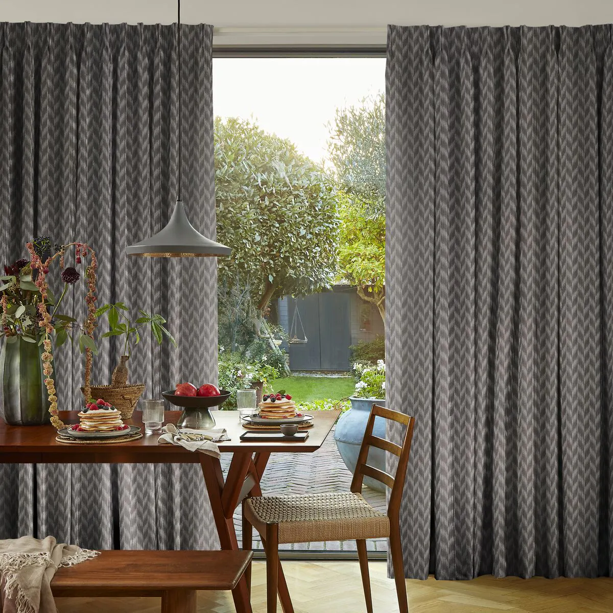 Dining area a wide glass window looking out to the garden, framed by grey patterned curtains, with a wooden table set beneath soft natural light.