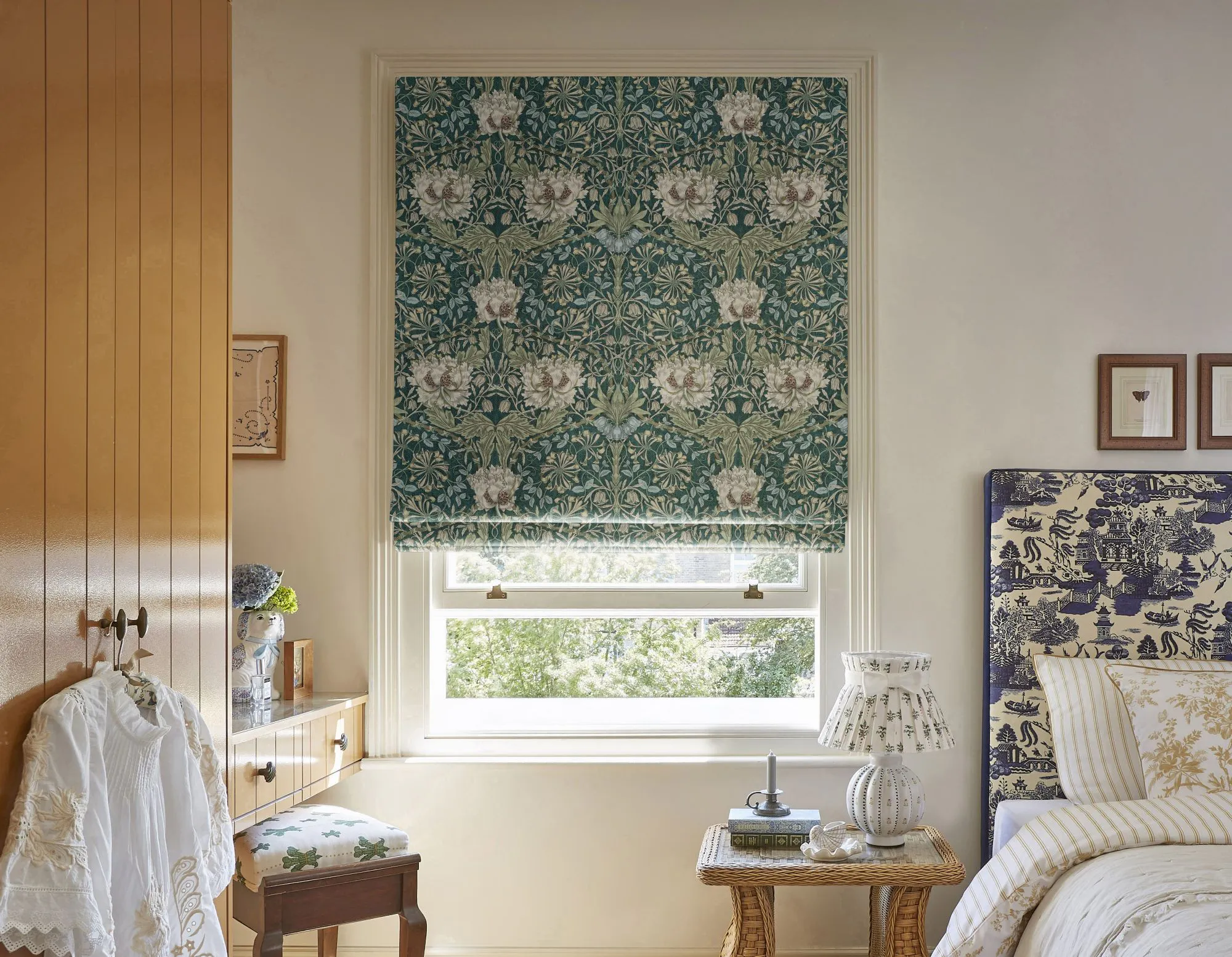 Cosy bedroom with a central window fitted with a patterned Roman blind, paired with layered textiles and classic wooden furniture.