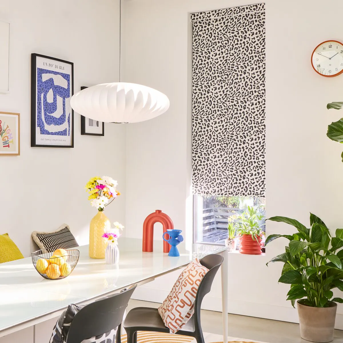 A bright, white-walled dining area with a monochrome leopard print Roller blind, styled with colourful wall art and accessories and leafy houseplants.