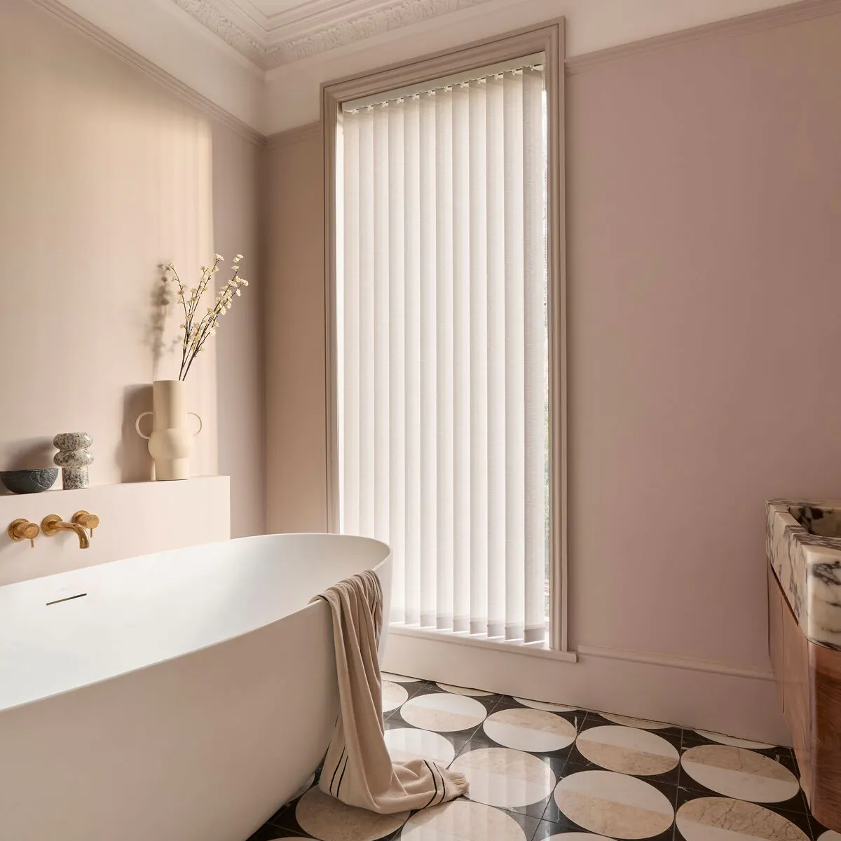 Tall window with white Vertical blinds diffusing light in a blush bathroom, beside a freestanding bath and patterned tiled floor.