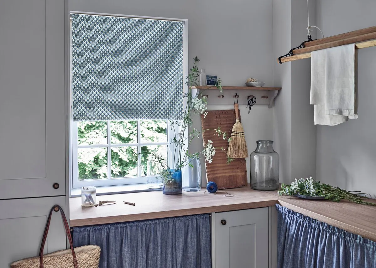 Blue patterned Roller blind over window in light utility room, with wooden shelf, glass vases, greenery, and folded linens on nearby surface.