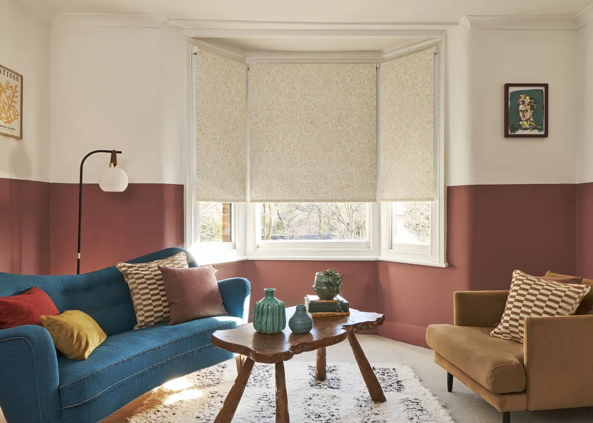 Bay window with three patterned Roller blinds partially lowered, soft light illuminating a cosy seating area with sofa, armchair and wooden table