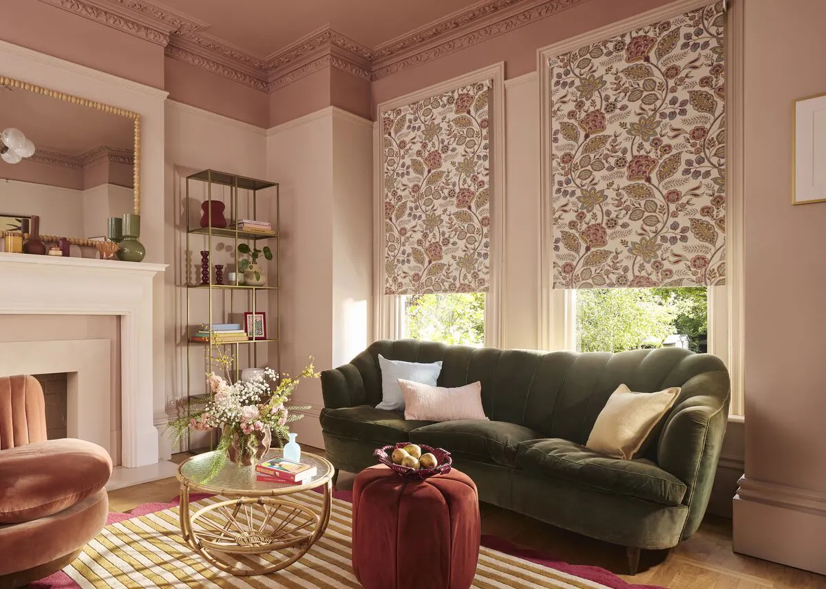Two tall windows with patterned Roller blinds partially lowered, casting warm light over a green sofa in a soft pink living room