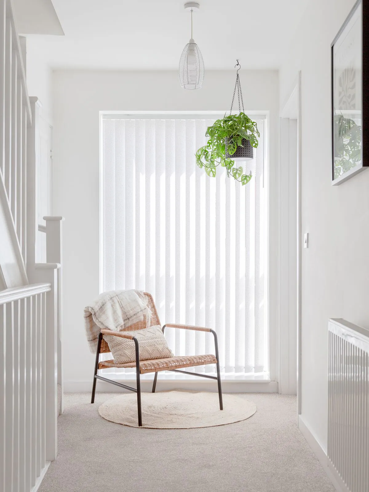 Bright hallway window with white Vertical blinds, paired with a wicker chair, cushion and hanging plant, creating a calm, airy corner.