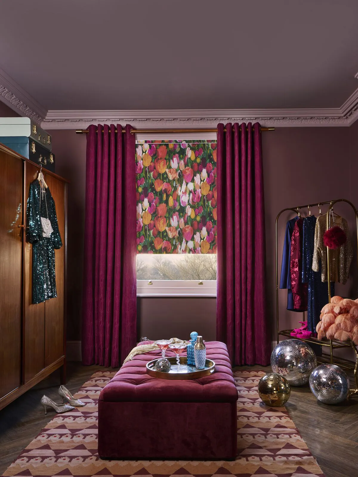 Window with vibrant floral blind partly raised, framed by plum curtains in a dressing room with clothes rail, mirror and metallic decor