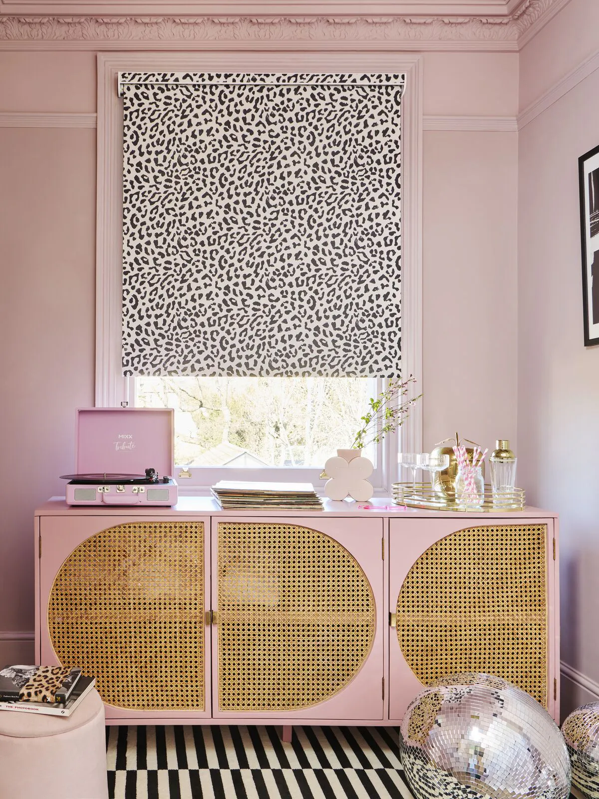 A colour-drenched pink dressing room featuring a monochrome leopard print Roller blind above a rattan-fronted sideboard and cluster of disco balls.