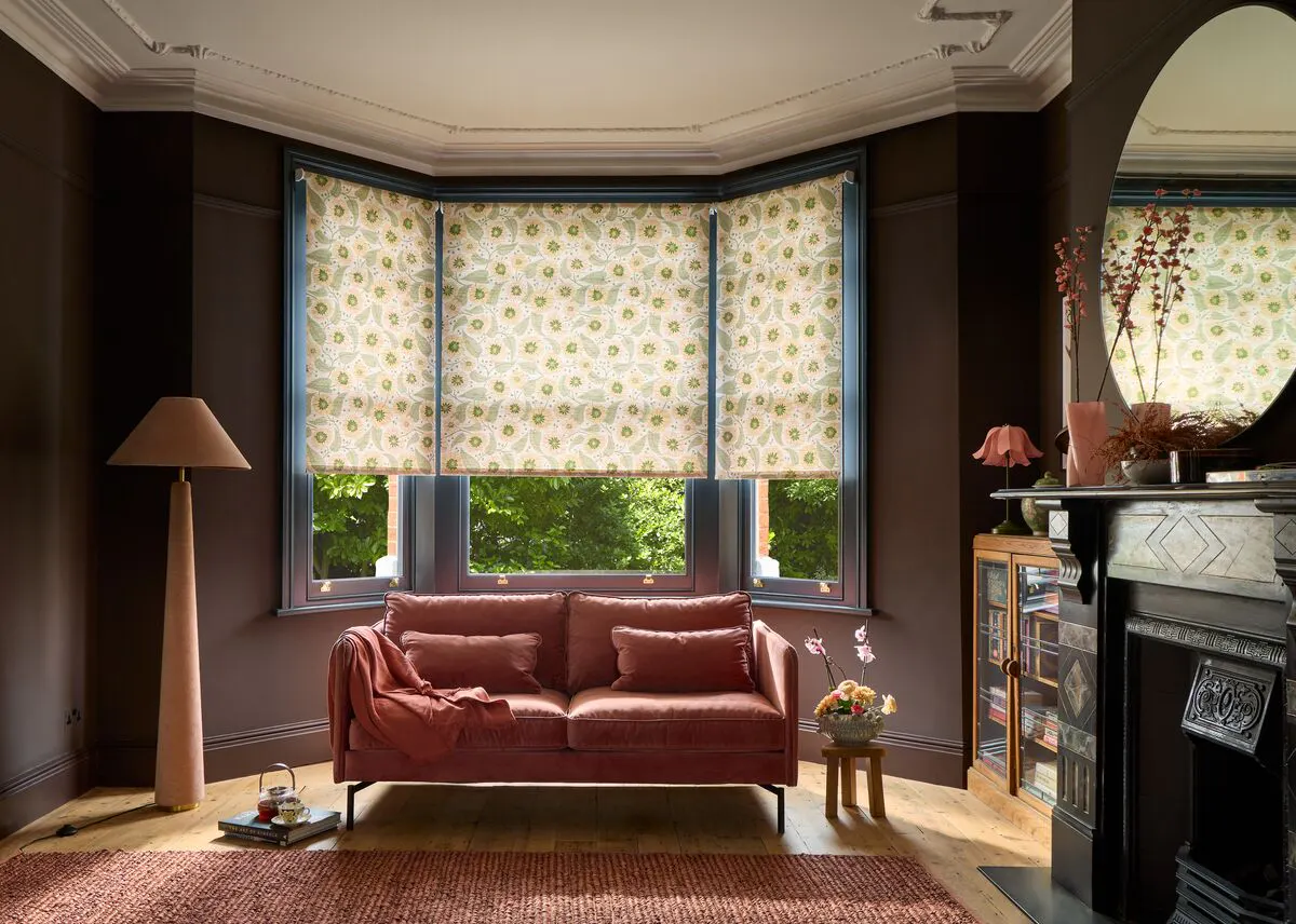 A Victorian-style living room featuring a dusky pink velvet sofa, ornate fireplace and dark walls framing a bay window with light patterned Roller blinds