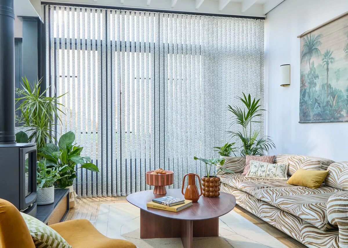 A bright living room with a low patterned sofa, curved wooden coffee table and several potted plants, all bathed in soft natural light filtering through floor-to-ceiling Vertical blinds.