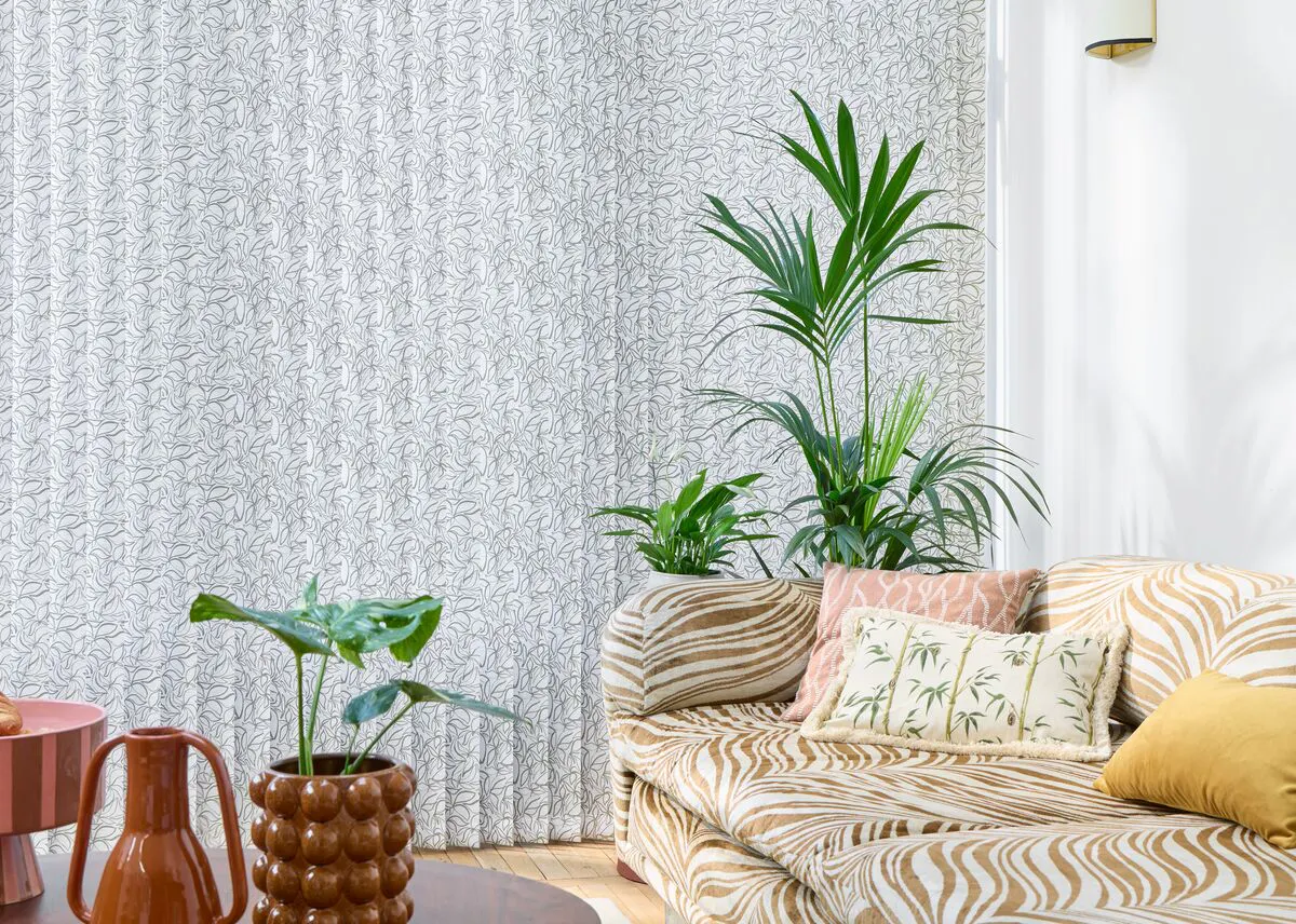 A bright living room corner with a low patterned sofa, cushions and several leafy houseplants in front of a subtly patterned full-height Vertical blind.